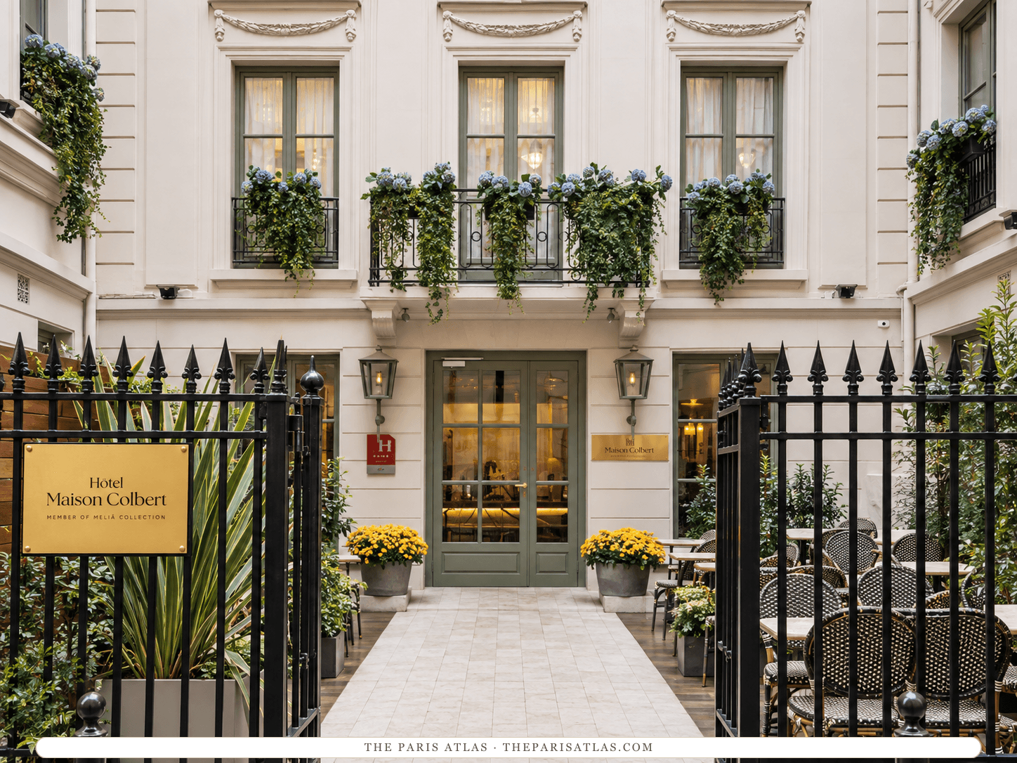 AI-generated illustration of the courtyard entrance to Maison Colbert: black iron railings, a gold sign reading Hôtel Maison Colbert, a stone path between café tables, yellow flowers in planters and green-framed glass doors beneath flower-filled window boxes.
