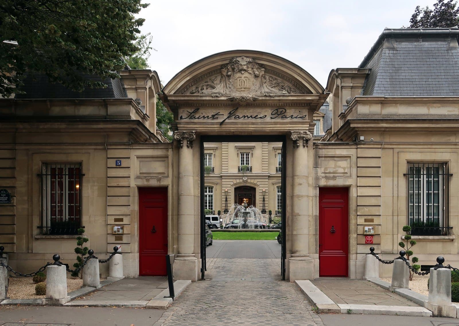 Photograph of the stone entrance gate of Saint James Paris, with a carved pediment, the words Saint James Paris in script, red doors in the pavilions on either side and the number 5, looking through to a fountain in the courtyard and the main façade beyond.